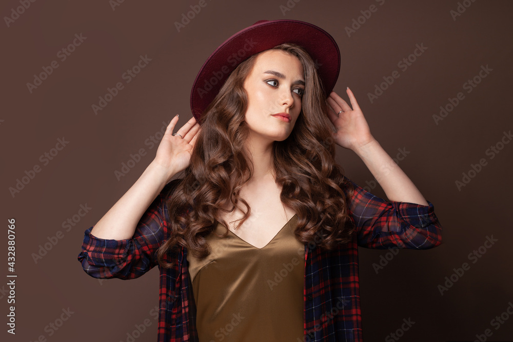Elegant woman in plaid shirt and fedora hat posing on brown background Elegant woman in plaid shirt and fedora hat posing on brown background