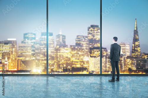Businessman standing and staring at the panoramic window with night city view in an empty big office room. Real estate and future success concept
