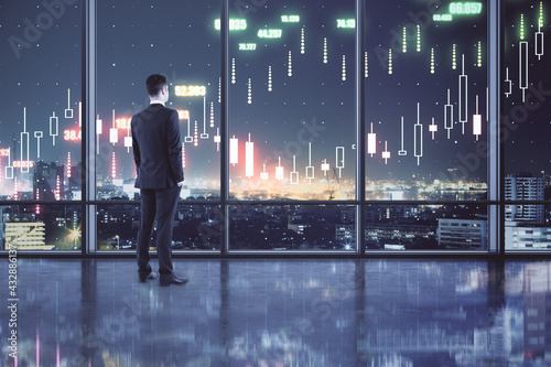 Profitable investing concept with buisnessman in spacious hall looking at night city and digital screen with financial chart candlestick. Double exposure.