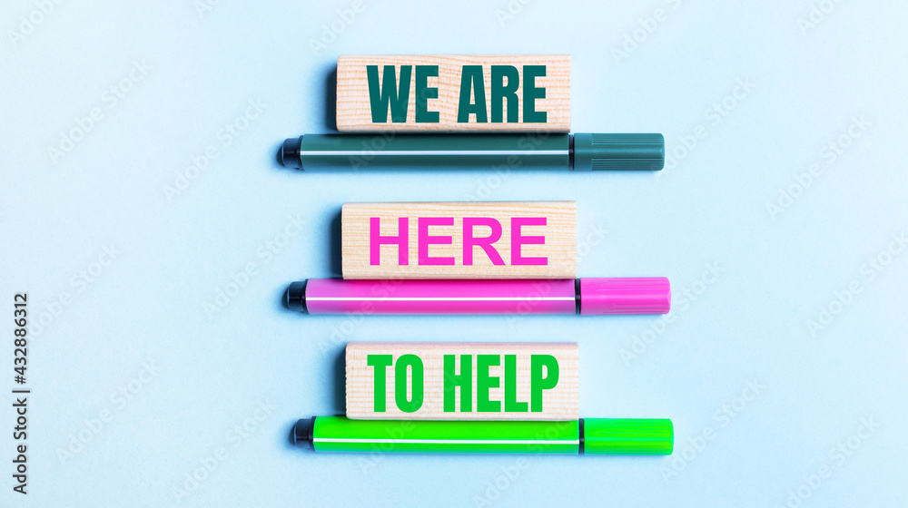 On a light blue background, there are three multi-colored felt-tip pens and wooden blocks with the WE ARE HERE TO HELP text. On a light blue background, there are three multi-colored felt-tip pens and wooden blocks with the WE ARE HERE TO HELP text.