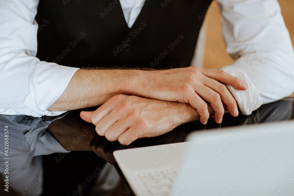 Crossed man's hands near white laptop placed on dark glass table. Focus is at hands. Business concept.