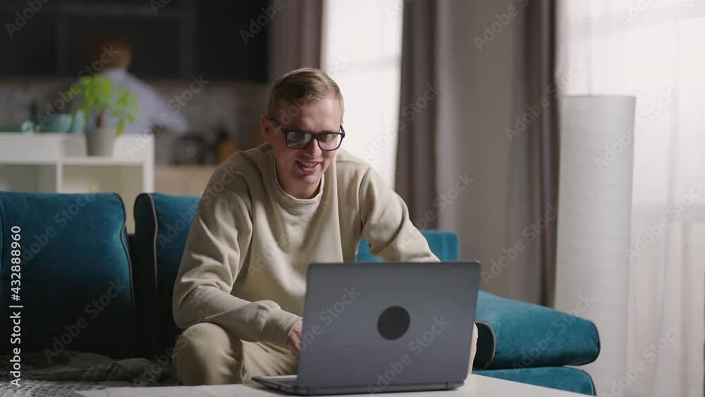 Man Sitting On Sofa At Home Making Video Call Using Laptop Computer on self isolation. Happy young blonde teacher man working from home, talking on laptop video conference.