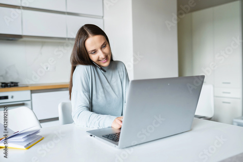 Busy multitask woman holds a smartphone with a shoulder and typing on the laptop keyboard, freelance woman using laptop for remote work from home, sits at the kitchen desk and has phone conversation © Vadim Pastuh