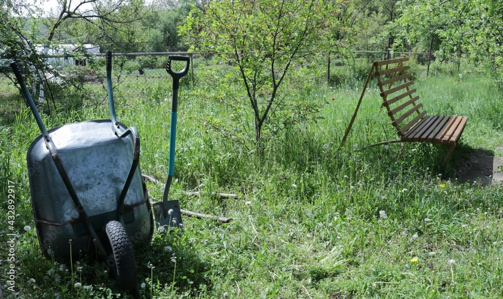 garden cart and shovel standing on a green lawn against the background of a spring garden and a wooden bench, a gardener's working tool among the grass, spring chores on the farm garden cart and shovel standing on a green lawn against the background of a spring garden and a wooden bench, a gardener's working tool among the grass, spring chores on the farm