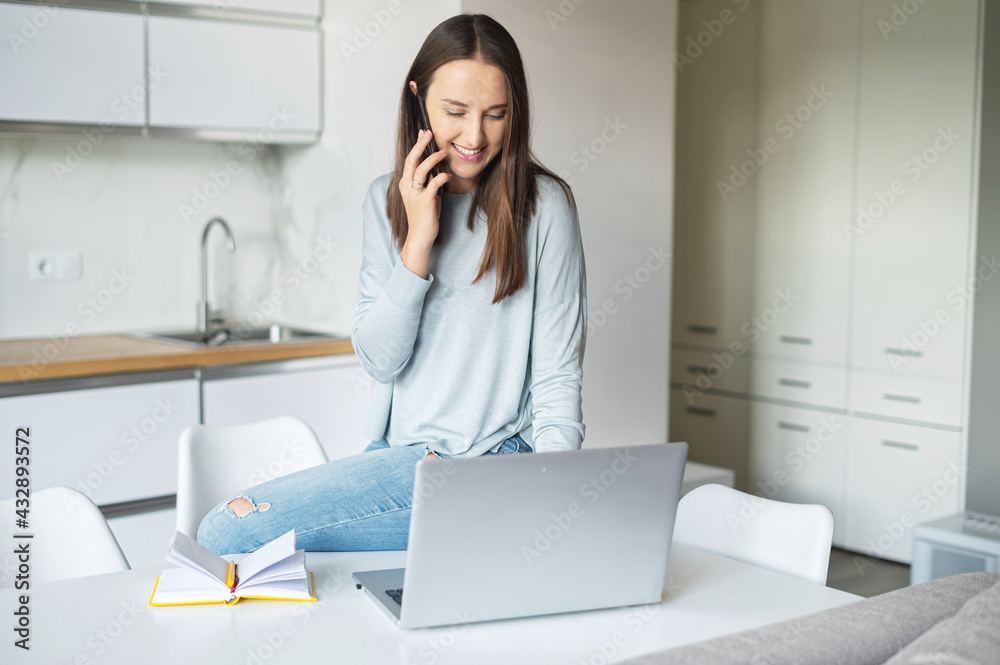 Charming young woman using laptop computer for remote work from home, talks on the smartphone leaned on the table in relaxed pose, female freelancer has pleasant phone conversation Charming young woman using laptop computer for remote work from home, talks on the smartphone leaned on the table in relaxed pose, female freelancer has pleasant phone conversation