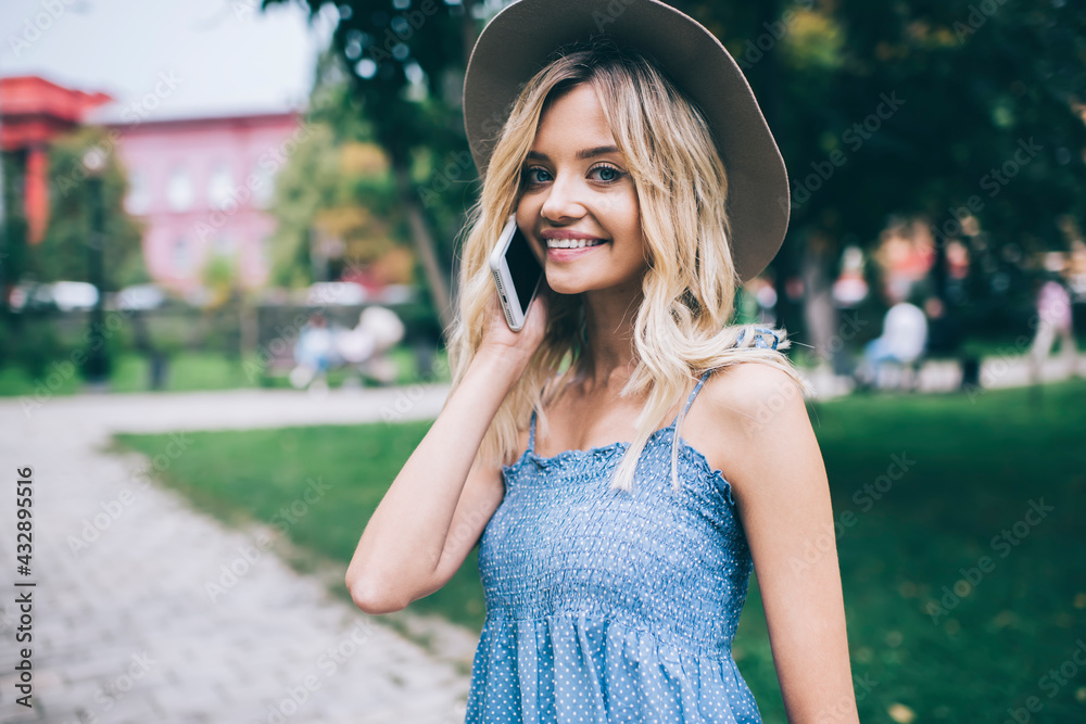 Half length portrait of cheerful female tourist smiling at camera while making friendly mobile conversation in roaming, happy hipster girl in trendy hat enjoying smartphone communication on leisure