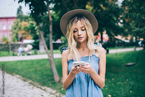 Millennial female blogger with cellphone device creating publication text for sharing to networks, beautiful hipster girl in trendy hat using modern smartphone gadget for web messaging on website