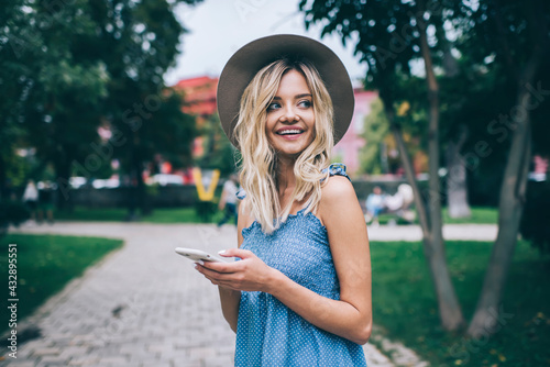 Funny hipster girl using cellular gadget for online networking during weekend leisure in park, happy Caucasian woman with mobile phone smiling and rejoicing during getaway vacations for travelling