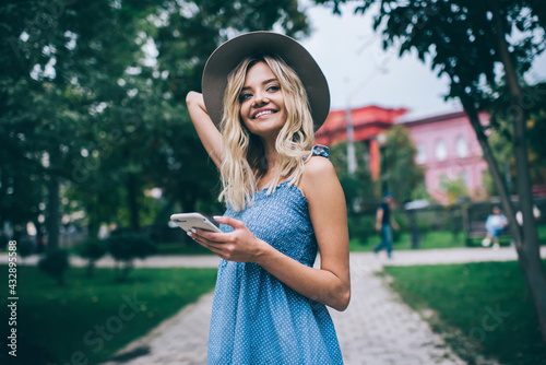 Joyful hipster girl using cellular gadget for online networking during weekend leisure in park, happy Caucasian woman with mobile phone smiling and rejoicing during getaway vacations for travelling