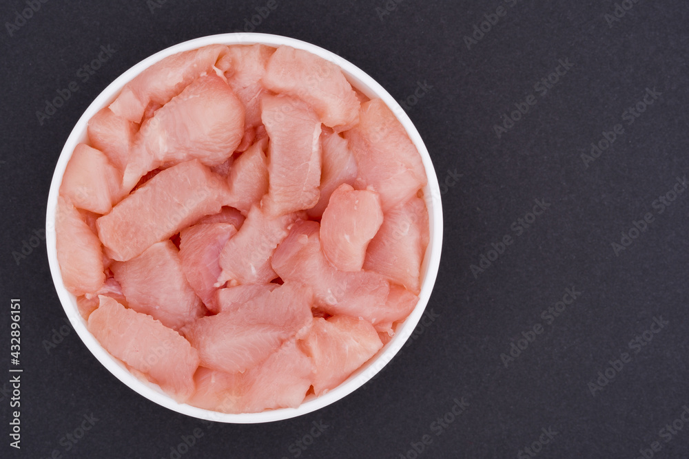 White glass bowl of sliced chicken fillet cubes. Isolated on a dark grey background. Top view close up White glass bowl of sliced chicken fillet cubes. Isolated on a dark grey background. Top view close up