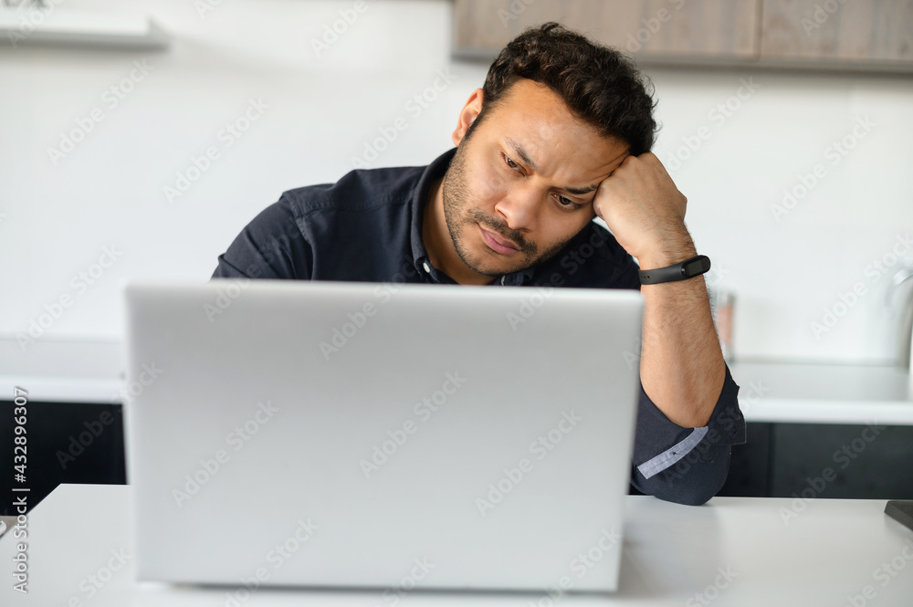 Upset multi-ethnic man is using laptop sitting at home alone, pensive young indian guy sits in the kitchen at with the laptop, rested head with hand, has financial problems and issues with project Upset multi-ethnic man is using laptop sitting at home alone, pensive young indian guy sits in the kitchen at with the laptop, rested head with hand, has financial problems and issues with project