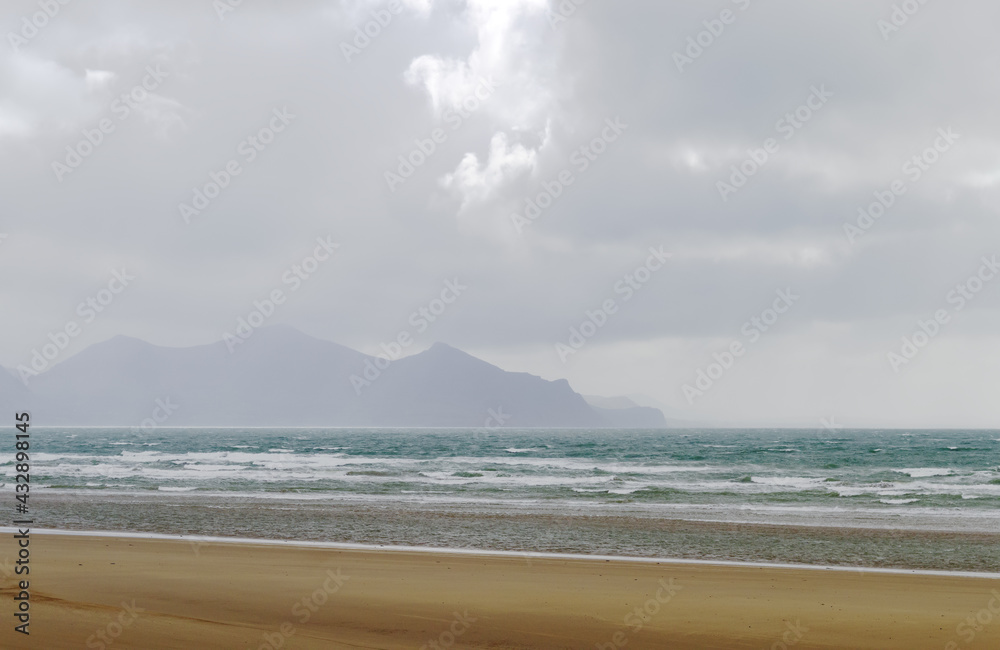 Llyn Peninsula, north Wales. Dramatic seaside landscape with stormy overcast skies. View to the distant mountains of the Llyn Peninsula on the horizon.  Windblown waves lap on a deserted beach.