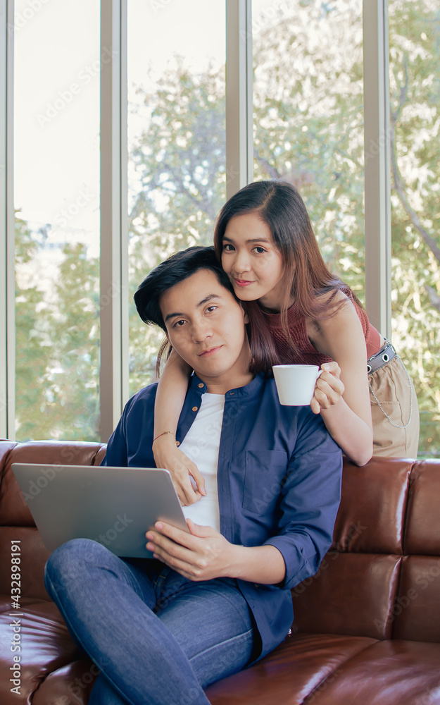 Asian beautiful woman smiling, holding cup of coffee, hugging, serving her boy friend while he using laptop for working in living room at home in morning, looking at camera. Lifestyle, Hobby Concept. Asian beautiful woman smiling, holding cup of coffee, hugging, serving her boy friend while he using laptop for working in living room at home in morning, looking at camera. Lifestyle, Hobby Concept.