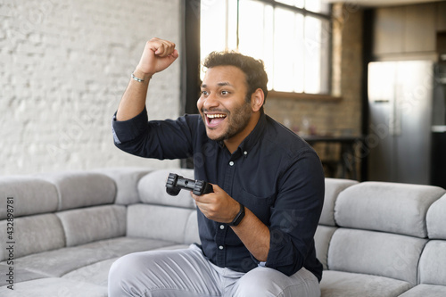 Excited mixed-race guy playing video game, indian young man sits on the couch, holds joystick, looks at camera and celebrate victory in game, latin man spends leisure at home, won game on the console