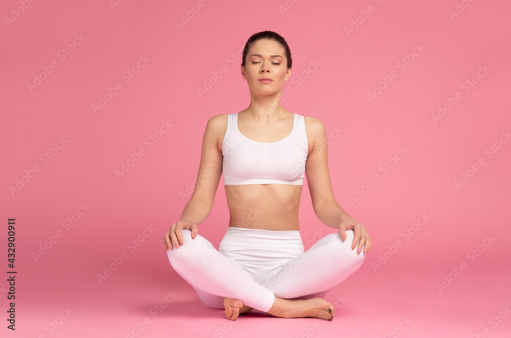 Young, beautiful woman in a white tracksuit practicing yoga meditation in the lotus position isolated on pink background Young, beautiful woman in a white tracksuit practicing yoga meditation in the lotus position isolated on pink background