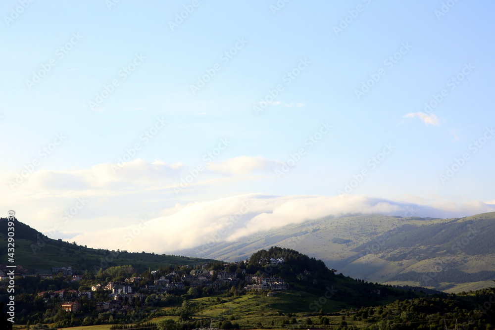 View of village perched on the mountains, among green trees, and other mountain topped by clouds in the background, Abruzzo, Italy