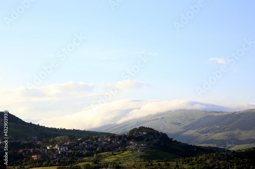 View of village perched on the mountains, among green trees, and other mountain topped by clouds in the background, Abruzzo, Italy © l