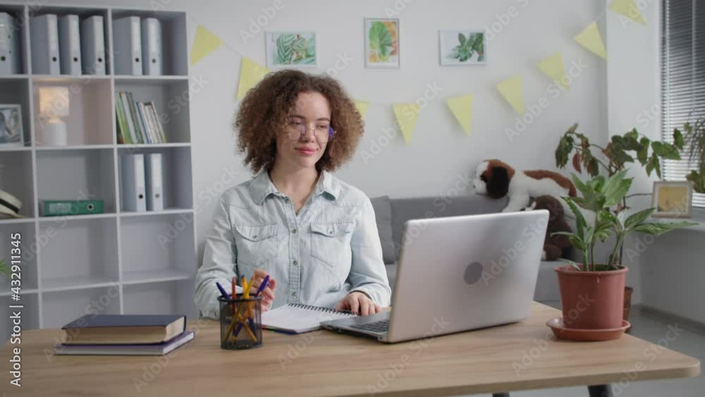 adult girl in glasses for vision teaches remotely lessons with teacher online via video communication on laptop writes in notebook smiling and looking at camera