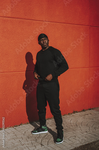 Confident Black Spanish male in black casual clothes and do-rag posing on a red wall background photo