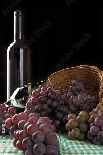Grapes, basket and overturned box with grapes falling on the table along with wine and a glass on a checkered tablecloth, black background, selective focus.
