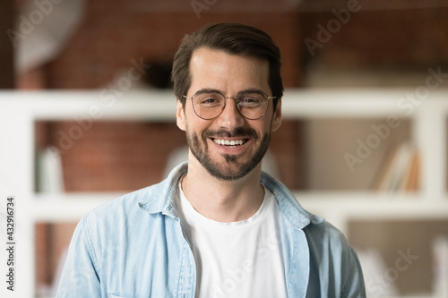 Head shot portrait confident smiling bearded businessman in glasses looking at camera, standing in office, successful happy young man employee entrepreneur posing for photo or recording video