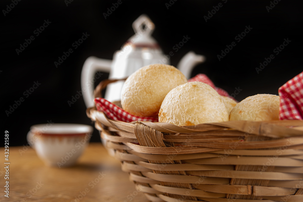 Brazilian cheese bread, checkered cloth basket with cheese breads on rustic wood with teapot and cup of coffee in the background, selective focus.