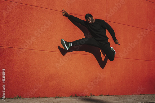 Cheerful Black Spanish male in black casual clothes and do-rag jumping against a red wall photo