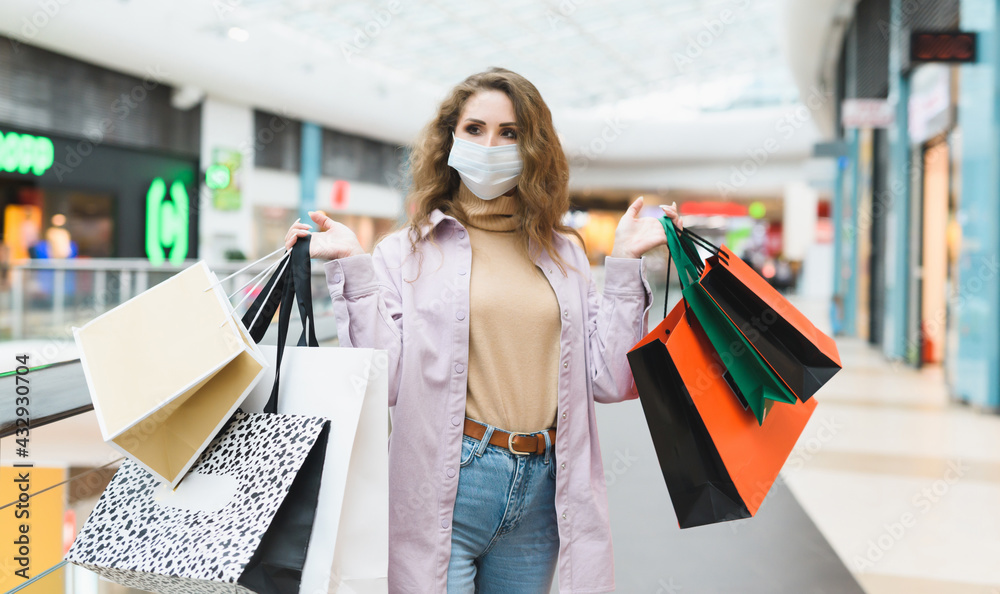 Portrait of a young woman shopaholic wearing medical mask in a shopping mall. A woman holds a lot of bags with clothes from different stores. Shopping during coronavirus Covid-19 lockdown Portrait of a young woman shopaholic wearing medical mask in a shopping mall. A woman holds a lot of bags with clothes from different stores. Shopping during coronavirus Covid-19 lockdown