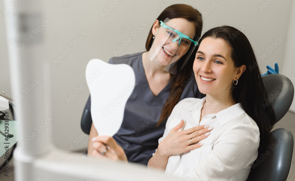 Satisfied patient! A young girl admires her smile after treatment, whitening or cleaning tooth enamel at the dentist. Kind friendly female doctor in the background. Dental clinic.