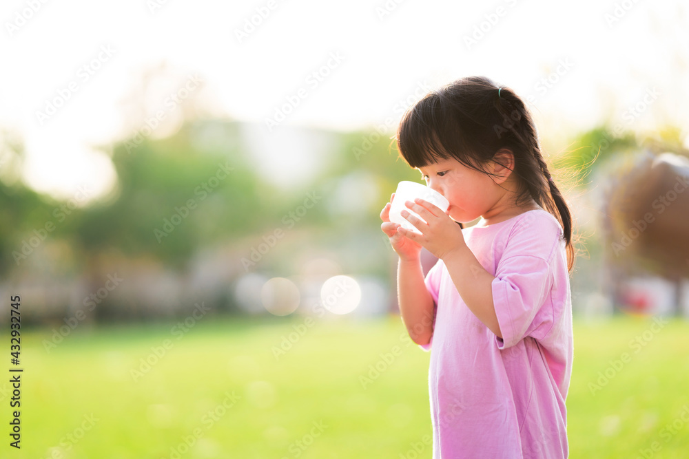 Selective focus. Cute 4 year old girl drinking some water from a plastic cup. Child is thirsty. Children protruding on the bright green lawn. Kid wore a pink shirt. Summer or spring. Selective focus. Cute 4 year old girl drinking some water from a plastic cup. Child is thirsty. Children protruding on the bright green lawn. Kid wore a pink shirt. Summer or spring.