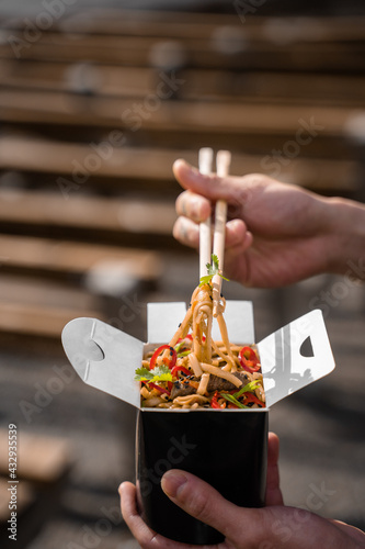 Fototapeta Naklejka Na Ścianę i Meble -  Wok in box in black food container. Holding spicy noodles with chopsticks. Fast food delivery service. Takeaway chinese street meal.