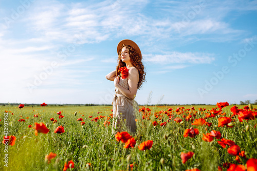 Beautiful girl posing in a poppy field. A young woman stands in a spring blooming garden. A curly-haired girl with poppies in her hands stands in a blooming field in summer. Sunny day summer landscape