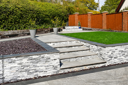 Steps from a patio to path and artificial grass in a residential back garden landscaped with light and dark grey porcelain paving slabs. No people. photo
