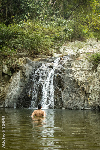 Hombre de espalda con el agua hasta la cintura caminando por un rio en medio de la naturaleza junto a una cascada