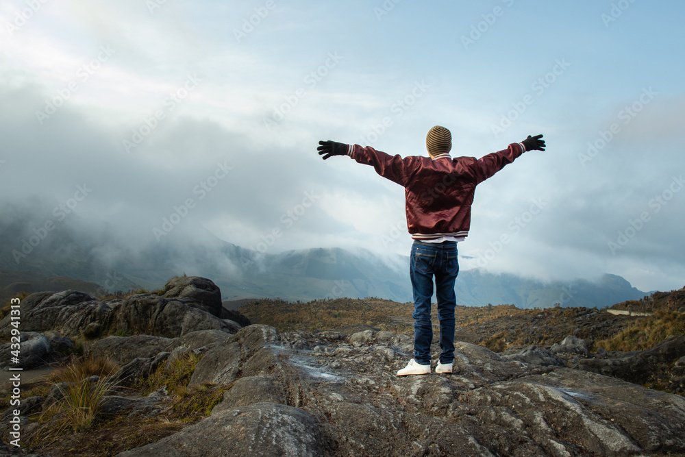 Hombre joven parado en la cima de una montaña con los brazos abiertos mirando el paisaje con la nubes azules de fondo Hombre joven parado en la cima de una montaña con los brazos abiertos mirando el paisaje con la nubes azules de fondo