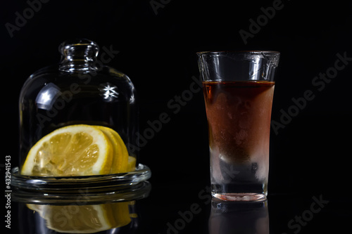 A glass of cognac on a black background next to a lemon in a container. Close-up