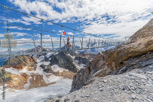 Woman on suspension bridge waving a Swiss flag. Titlis cliff walk with glacier in the Uri Alps. Viewpoint at 3028 meters in Summer season, Bern canton. The highest suspension bridge in Europe. photo