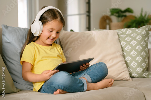Smiling little girl excited about what she sees on screen of digital tablet during distance education, sitting on sofa with pillows. Children and gadgets concept Joyful learning process.