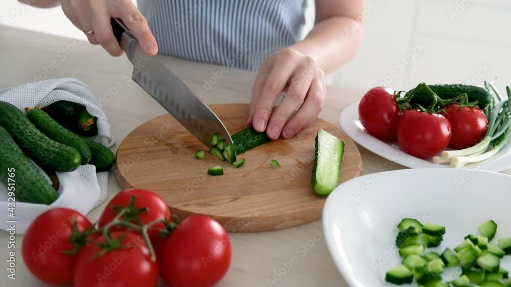 Woman cutting cucumber for salad on wooden cutting board on kitchen table with cucumbers, tomatoes branch and scallion around. Cooking dieting salad full of vitamins and antioxidants. Woman cutting cucumber for salad on wooden cutting board on kitchen table with cucumbers, tomatoes branch and scallion around. Cooking dieting salad full of vitamins and antioxidants.
