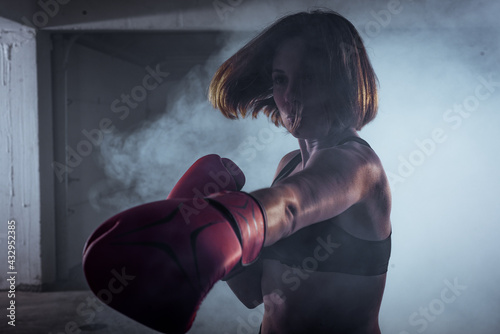 Sporty girl with a competitive fight attitude doing boxing exercises and making a direct hit on smoky background