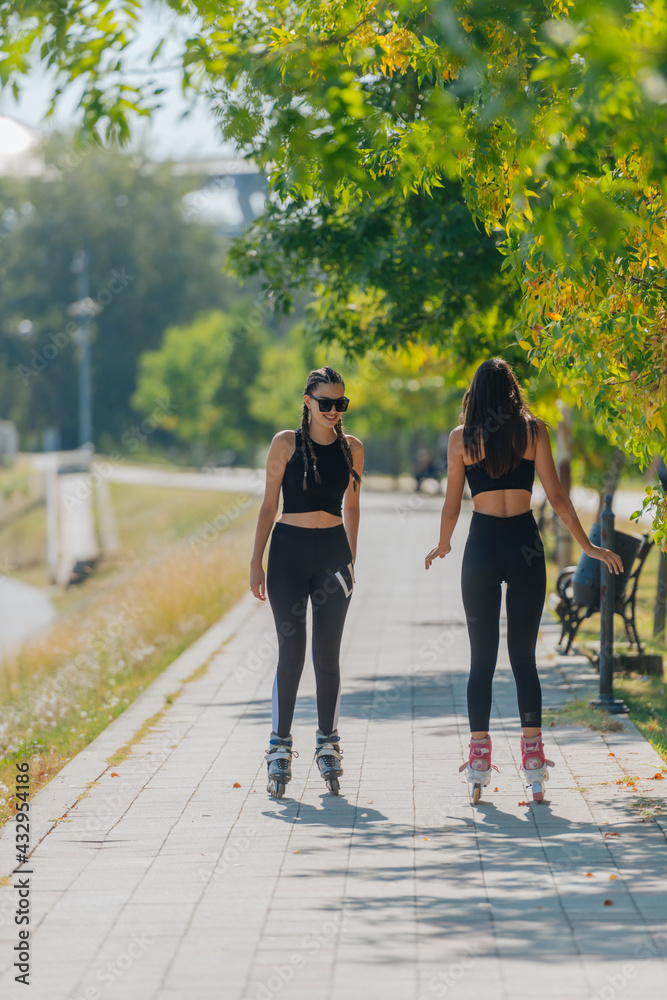 Obraz premium Rearview of two young fit women on roller skates riding outdoors on urban street in the park.