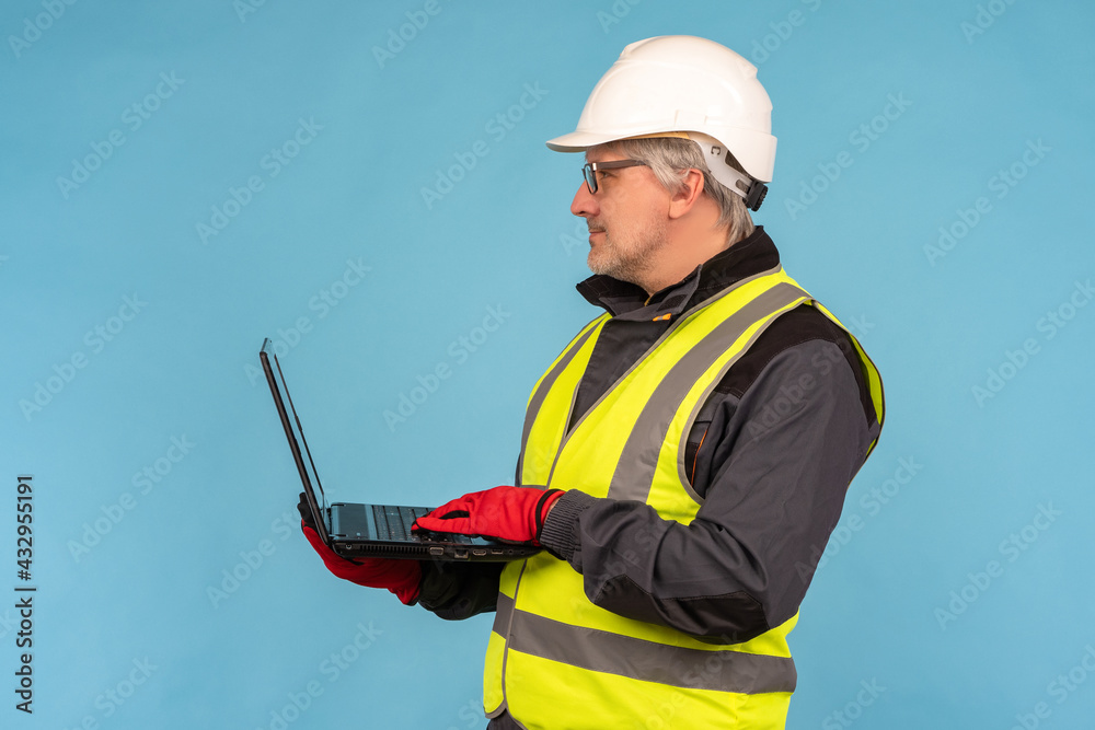 Civil engineer with a laptop in his hands. A man in an orange vest and hard hat. An architect with a laptop stands sideways to the viewer. A man in work clothes on a blue background. Civil engineer with a laptop in his hands. A man in an orange vest and hard hat. An architect with a laptop stands sideways to the viewer. A man in work clothes on a blue background.