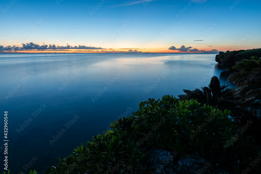 Long exposure scene of peaceful sea landscape. Vegetation on top of cliff. Horizon with a soft light, orange and yellow. Blue ocean with a smooth surface.