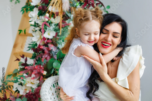 A woman with a little girl in beautiful white dresses in a bright studio decorated with flowers. Mother and daughter play, hug, have fun. © Elena 