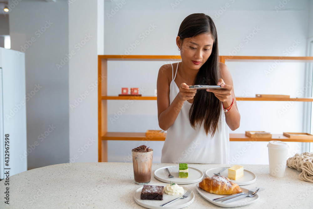 Asian woman using smartphone taking a photo of tasty cake dessert on white plate on the table. Female blogger influencer photographing sweet bakery and beverage for review in food restaurant blog Asian woman using smartphone taking a photo of tasty cake dessert on white plate on the table. Female blogger influencer photographing sweet bakery and beverage for review in food restaurant blog