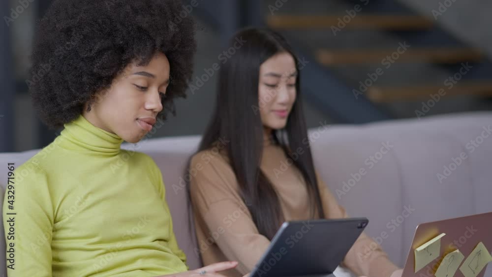 Side angle view of smiling charming African American woman messaging online on tablet with blurred Asian roommate typing on laptop keyboard at background. Remote working in home office Side angle view of smiling charming African American woman messaging online on tablet with blurred Asian roommate typing on laptop keyboard at background. Remote working in home office
