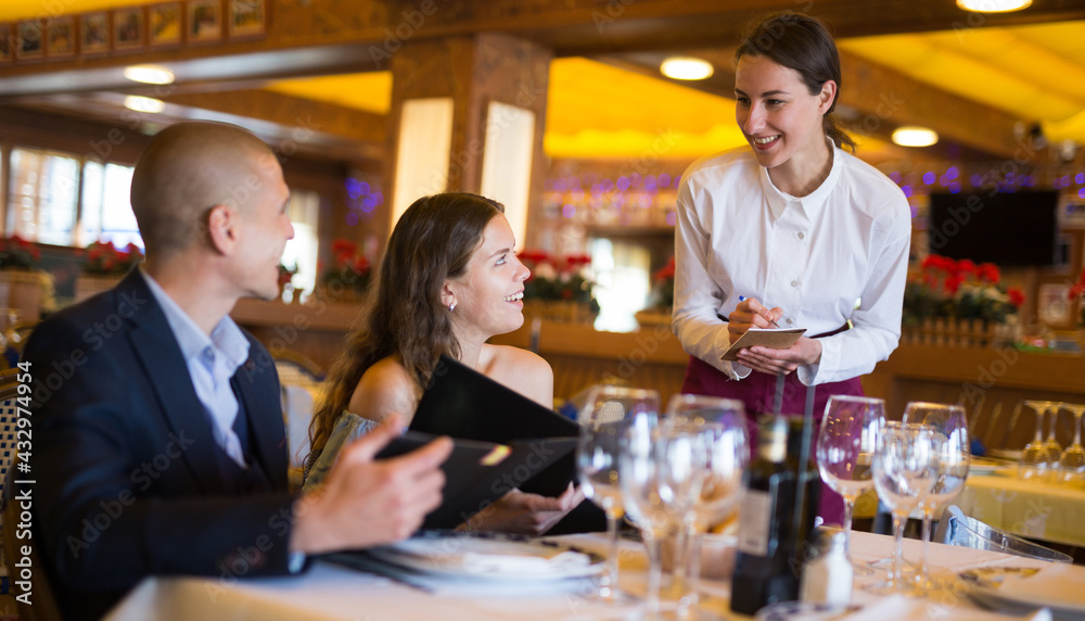 Attractive smiling woman waiter receiving order from guest in fashionable restaurant