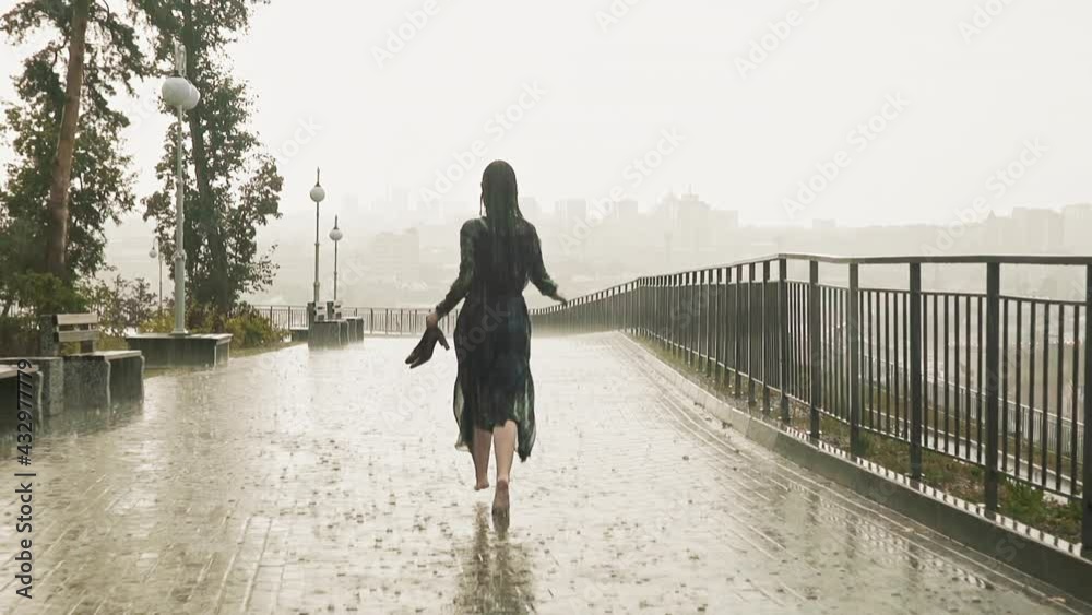 Graceful long haired woman in wet dress runs holding heeled shoes along paved promenade under heavy rain on summer day backside view slow motion