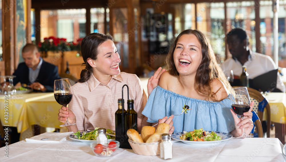 Two girls are discussing something fun at a table in a restaurant - obrazy, fototapety, plakaty 