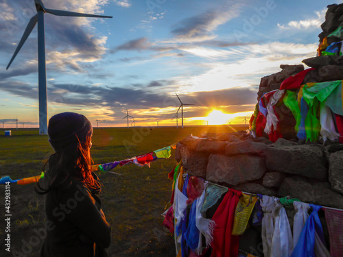 A woman in beanie walking around a Heaps of stones (Aobao) build on a vast pasture in Xilinhot in Inner Mongolia. The Heaps has colorful prayer flags attached to it. The sun is setting. Wind turbines